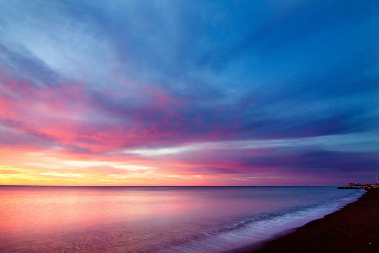 Pink and blue sunset over a calm sea, waves reaching a dark beach.
